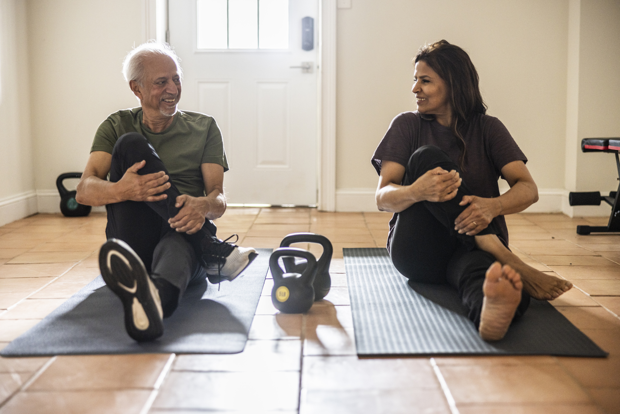 Senior man and woman sit on exercise mats performing a glute stretch, with kettlebells on the floor between them