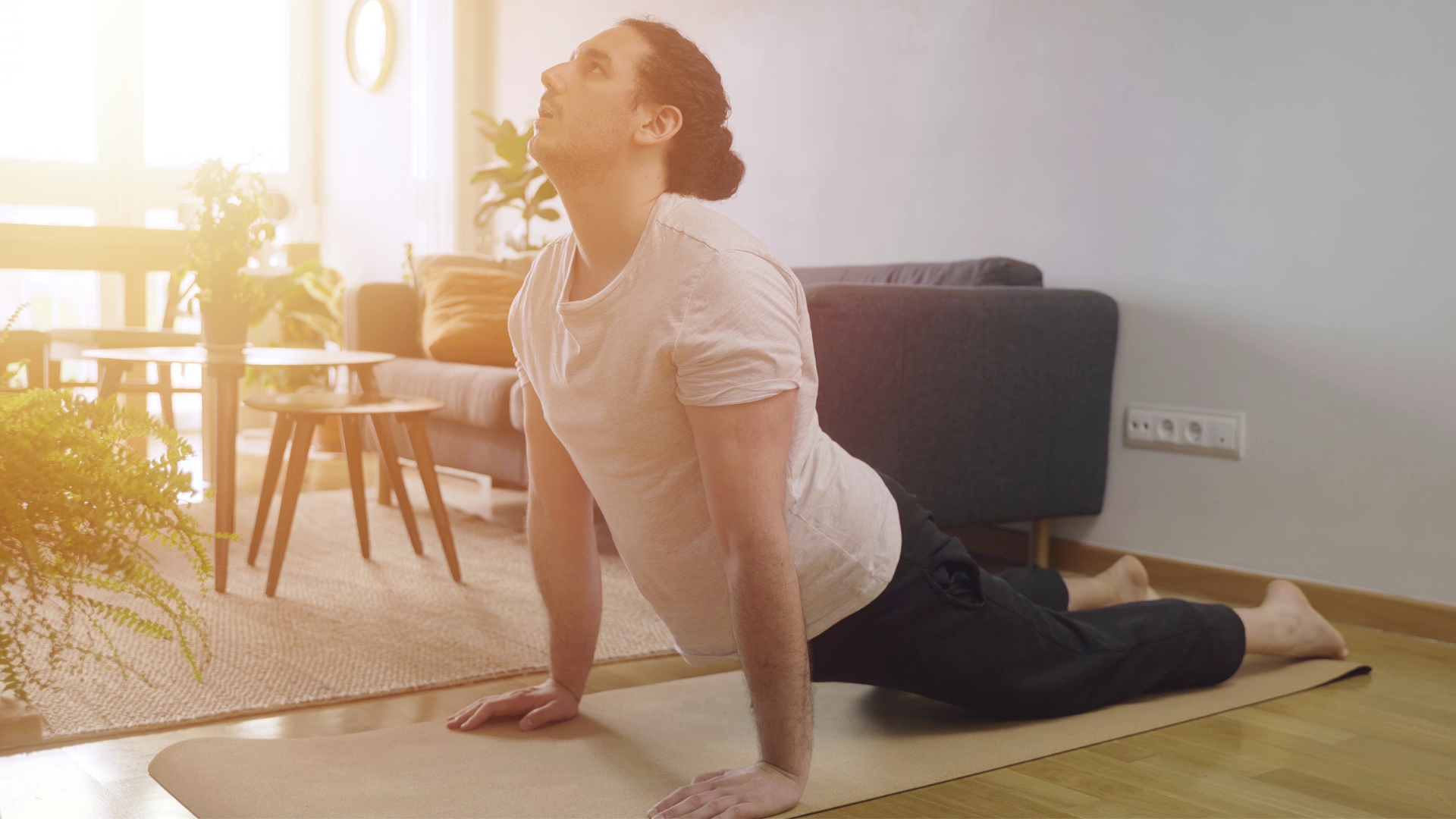 man wearing a white tshirt performing an extended spinal stretch from an exercise mat on the floor. he's sideways to the camera and there's living room furniture behind him with sunlight shining through.