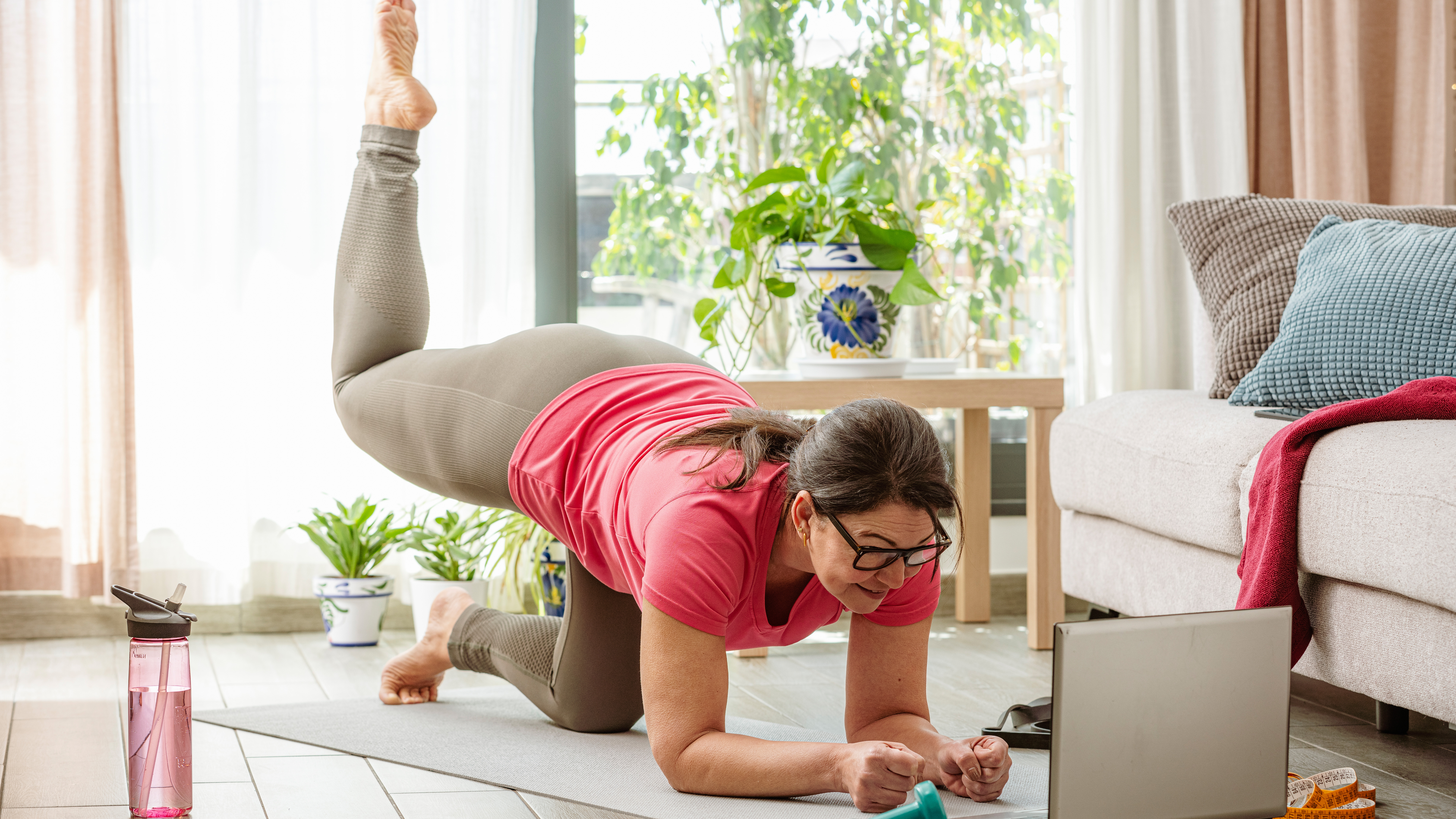 A woman practices Pilates at home in her living room on a mat. She is balancing, close to the ground, on her forearms and her bent left knee. Her right leg is bent at 90° and elevated behind her. In the room we see a wtaer bottle, a couch, a plant and a coffee table. A laptop is open in front of her.