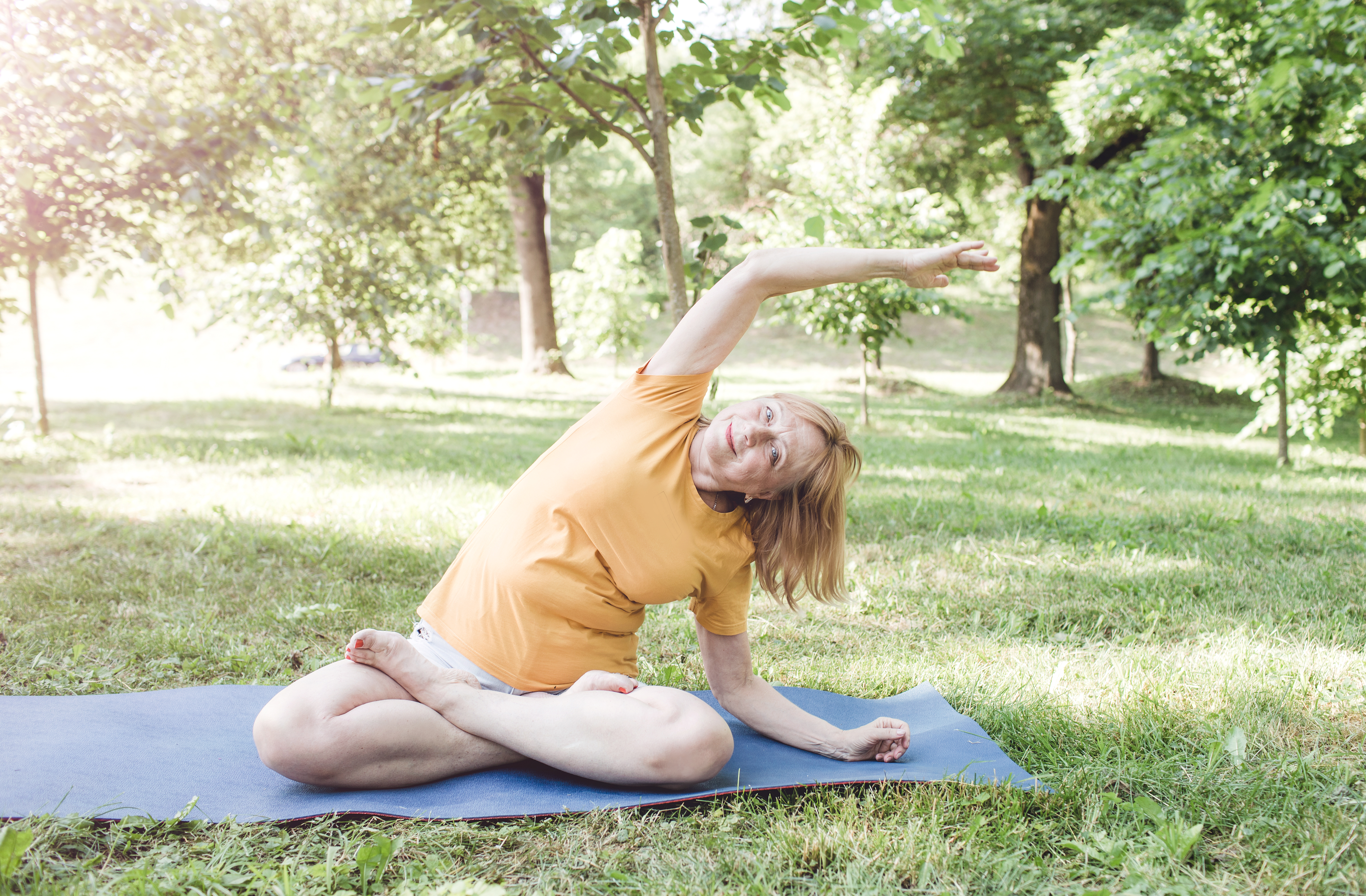 A woman sits in a park on a yoga mat, with her legs crossed, her left forearm on the mat next to her and her right arm reaching up and across. She is smiling and we see leafy trees and grass behind.