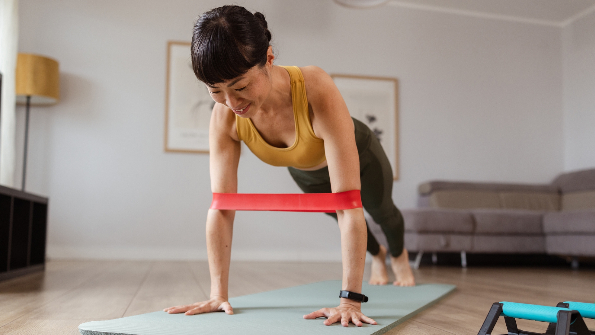 woman in a high plank position with a short resistance band around her arms around the elbows. she's on an exercise mat in a living room