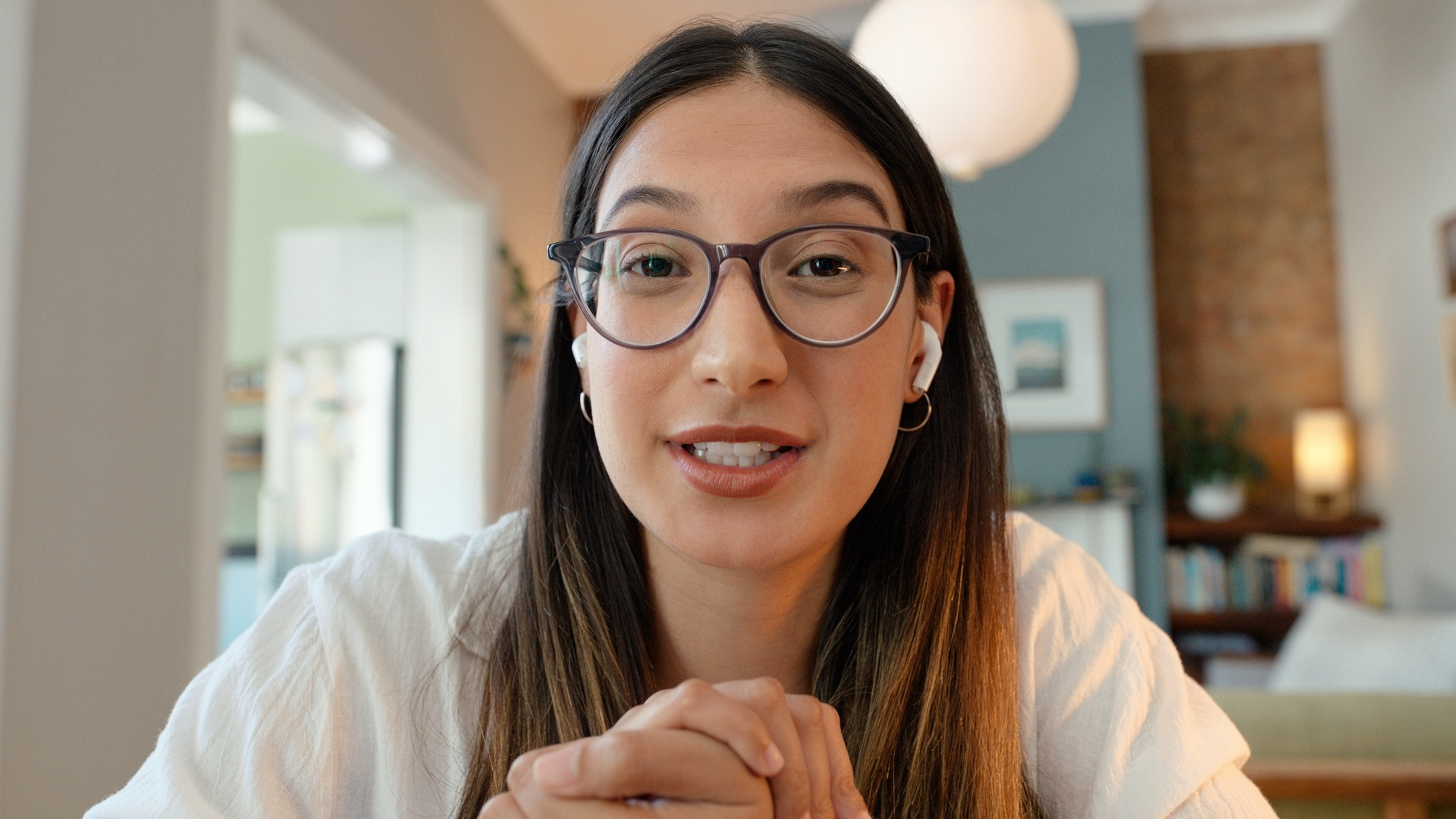 Woman wearing glasses and white earbuds in domestic setting looks at viewer
